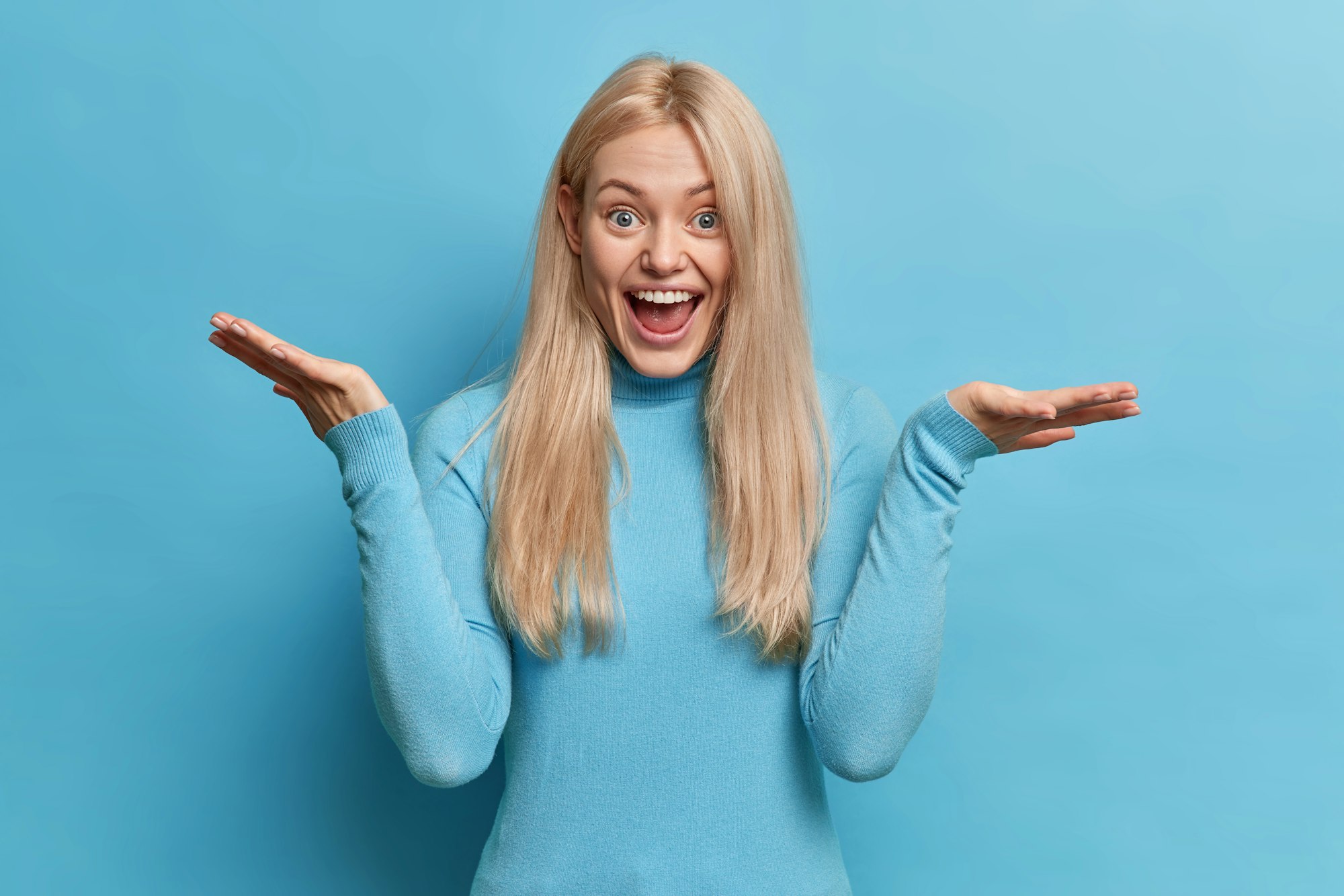 Happy unaware Caucasian woman with blonde hair spreads palms shrugs shoulders and has doubtful expre